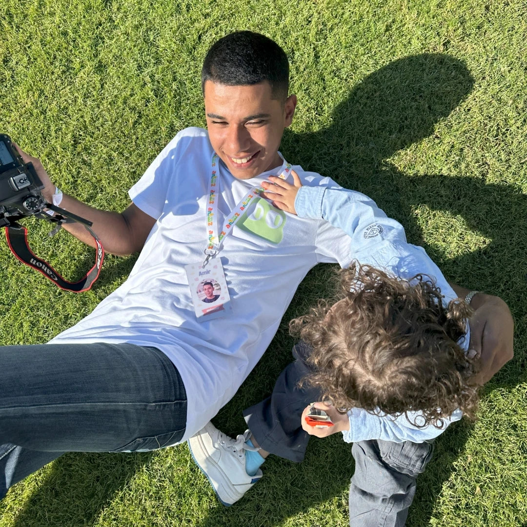 A joyful moment captured outdoors as a young man, Amir the founder of Antooz, enjoys playtime with a child, camera in hand, on lush green grass.